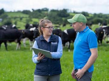 Lakeland Dairies team member, Susan Casey speaking to a Milk Supplier about his farm. Cows graze in the background.