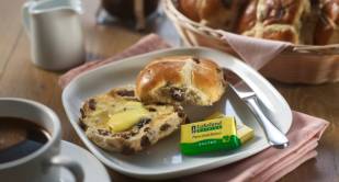 Hot cross bun, cut in half, with melted butter on, next to two packs of Lakeland Dairies Salted Butter portions