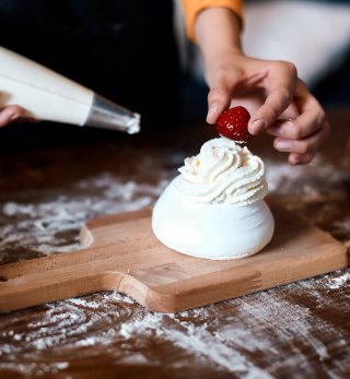 Serving suggestion recipe of a meringue, with Lakeland Dairies Whipped Cream piped on top, with a close up of chef's hand placing a fresh strawberry on top and holding a piping bag of cream in the other hand